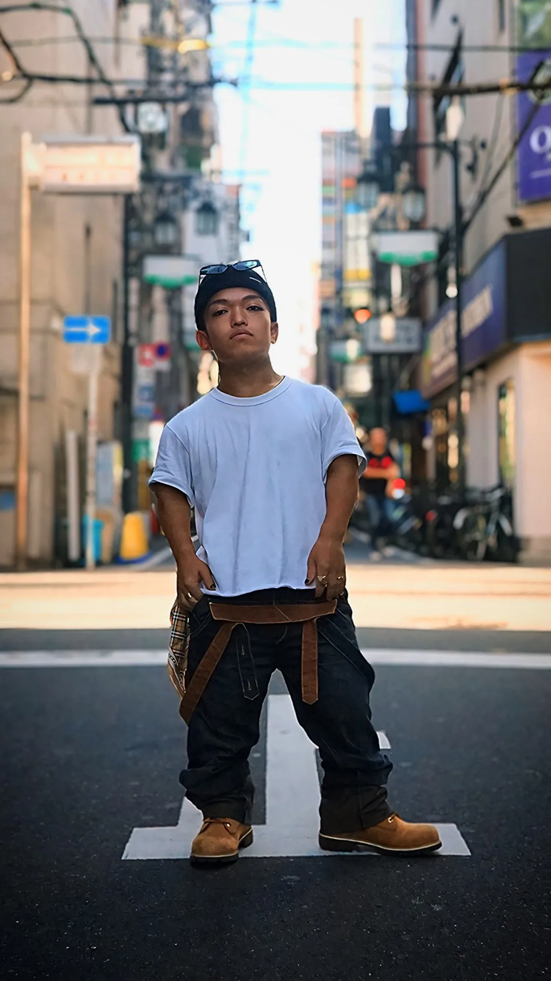 街の交差点に立つ、小人症のKRUMPダンサーDWARF。白いTシャツにニット帽、ブーツ姿でカメラを見据えている